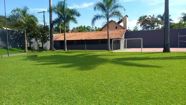 Campo gramado com traves de futebol, sede ao fundo entre palmeiras
